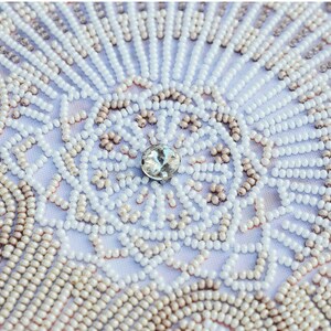 May include: Close-up of a white fabric with a beaded design. The beads are arranged in a circular pattern with a clear rhinestone in the centre.