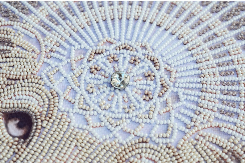 May include: Close-up of a white fabric with a beaded design. The beads are arranged in a circular pattern with a clear rhinestone in the centre.