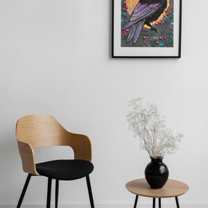 Puede incluir: Una obra de arte enmarcada que presenta un cuervo negro con plumas azules iridiscentes posado en una rama con bayas rojas y una luna amarilla en el fondo.