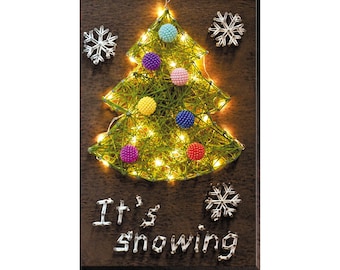May include: A string art Christmas tree with lights and colorful ornaments. The tree is on a brown wooden background with the words "It's snowing" written below it.