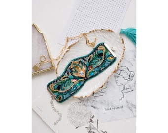 May include: A beaded bracelet with a floral design in shades of blue, green, and gold. The bracelet is on a white ceramic dish with a gold rim.
