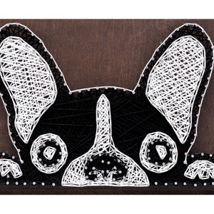 May include: A black and white string art piece depicting a French bulldog with its ears perked up and its head peeking out from behind a brown wooden frame.
