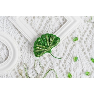 Könnte beinhalten: Eine grüne Ginkgoblatt-Brosche aus Perlen. Die Brosche liegt auf einem weißen gestrickten Stoffhintergrund.