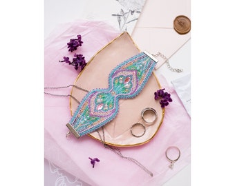 Puede incluir: Un brazalete de cuentas con un diseño floral turquesa, rosa y verde. El brazalete está sobre un plato rosa y dorado con una cadena plateada.