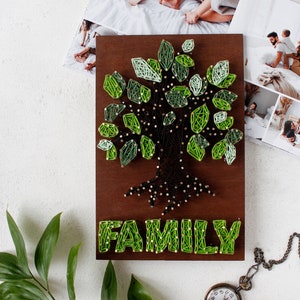 Peut inclure: Une décoration murale en bois avec un arbre en fil vert et le mot "FAMILY" en dessous. L'arbre a un tronc et des racines marrons.