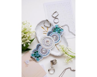 Puede incluir: Un brazalete de cuentas blancas y azules con cuentas verdes y transparentes. El brazalete está en un plato de cerámica blanca con un diseño de hoja de oro. El brazalete tiene un cierre plateado.