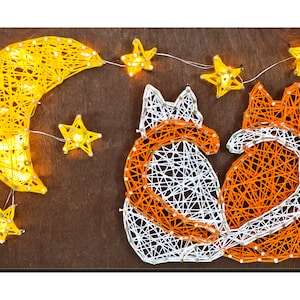 Puede incluir: Arte de hilo de dos zorros con sus colas entrelazadas, rodeados de una luna creciente y estrellas. Los zorros están hechos con hilo blanco y naranja, mientras que la luna y las estrellas son amarillas.