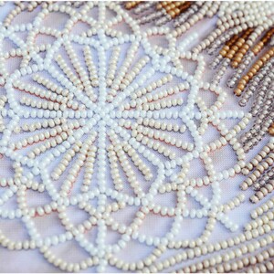 May include: Close-up of a white fabric with a beaded design. The beads are arranged in a circular pattern, creating a mandala-like design. The beads are white, cream, and gold.