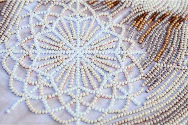 May include: Close-up of a white fabric with a beaded design. The beads are arranged in a circular pattern, creating a mandala-like design. The beads are white, cream, and gold.