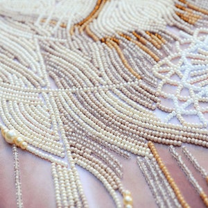 May include: Close-up of a beaded fabric with a white, cream, and gold pattern. The beads are arranged in a geometric design, creating a textured and intricate surface.