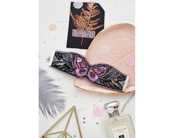 May include: A beaded bracelet with a purple butterfly design. The bracelet is on a pink heart-shaped dish with a silver clasp.