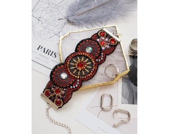 May include: A beaded bracelet with a black and red design featuring a circular pattern with gold accents. The bracelet has a silver clasp and chain.
