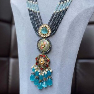 May include: A multi-strand necklace with blue and white beads and a large pendant with a floral design. The pendant is made of gold and has a blue stone in the center.