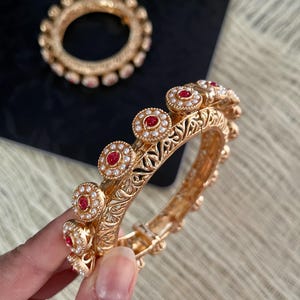 May include: Two gold bangles with intricate designs and red and white stones. The bangles are stacked on top of each other.