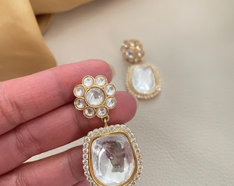 May include: Gold dangle earrings with a large clear crystal surrounded by smaller crystals. The top of the earring features a cluster of smaller crystals in a flower shape.