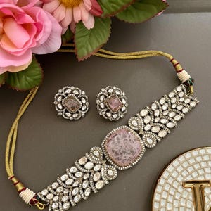 May include: A silver-toned necklace and earring set with a pink center stone and clear accent stones. The necklace has a gold-colored cord. Pink flowers are in the background.