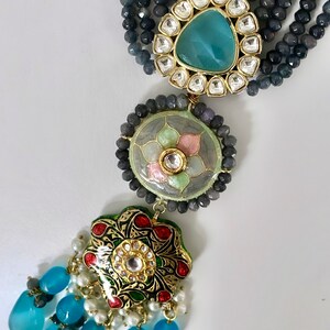 May include: A multi-strand necklace with gray beads and a pendant featuring a blue stone surrounded by smaller white stones. The pendant is attached to a round, green and pink enamel pendant with a floral design. The necklace ends with a tassel of blue teardrop-shaped stones.