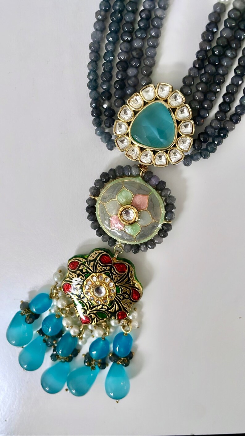 May include: A multi-strand necklace with gray beads and a pendant featuring a blue stone surrounded by smaller white stones. The pendant is attached to a round, green and pink enamel pendant with a floral design. The necklace ends with a tassel of blue teardrop-shaped stones.