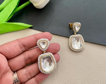 May include: A pair of gold earrings with clear, faceted gemstones. The earrings have a teardrop shape and are accented with small, clear stones.
