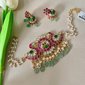 May include: A gold necklace with a pink and green floral design, featuring white pearls and green stones. The necklace is accompanied by a pair of matching earrings.