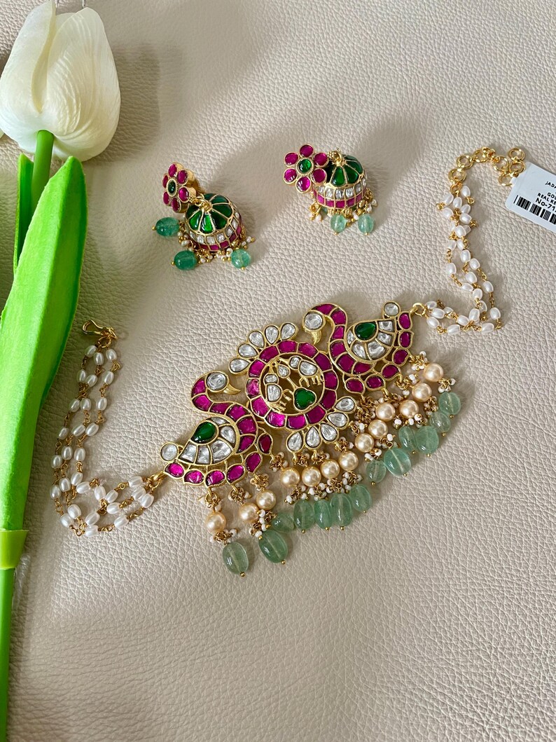 May include: A gold necklace with a pink and green floral design, featuring white pearls and green stones. The necklace is accompanied by a pair of matching earrings.