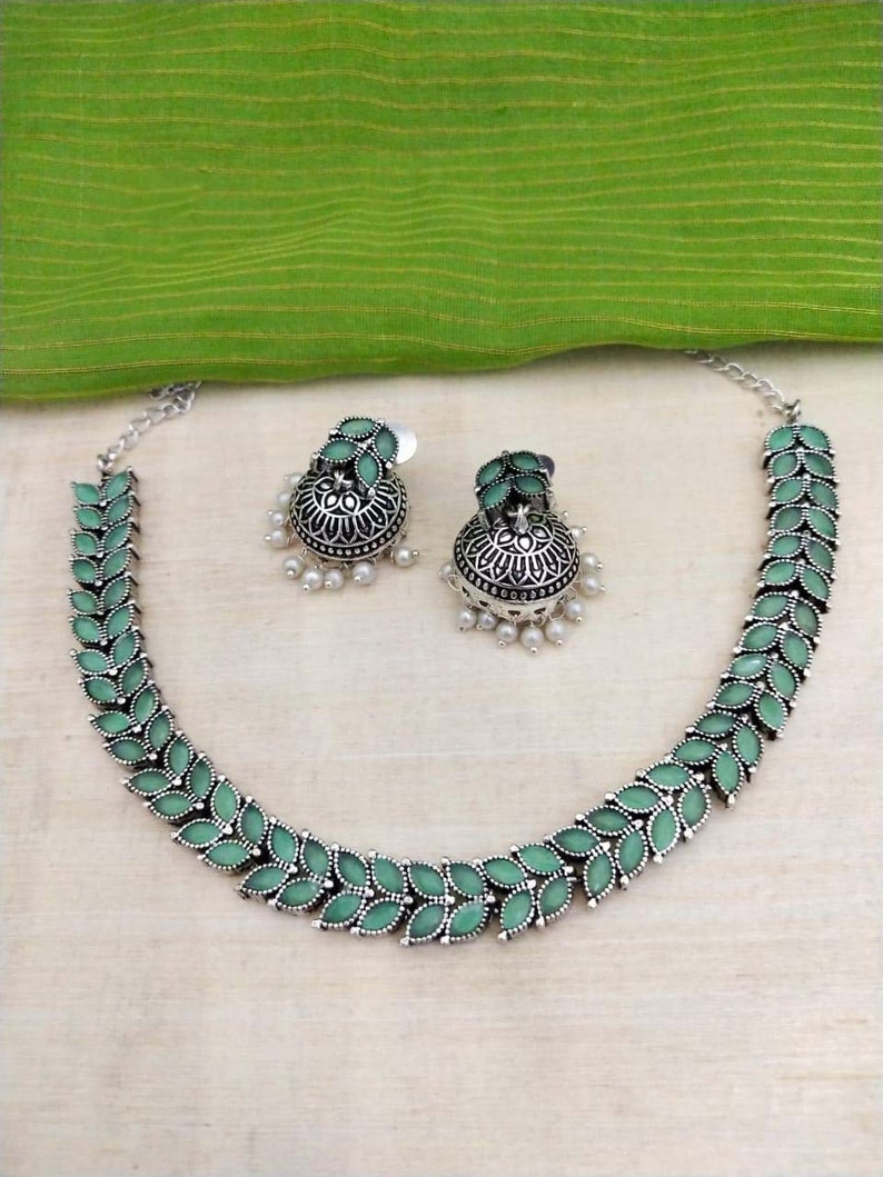 May include: A silver necklace with green leaf-shaped pendants and matching earrings. The earrings are jhumka style with a pearl drop.
