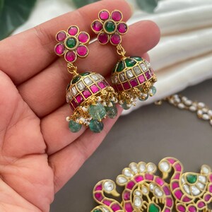 May include: A gold necklace with a pendant featuring two peacocks with pink and green gemstones. The necklace is paired with matching earrings with pink and green gemstones.