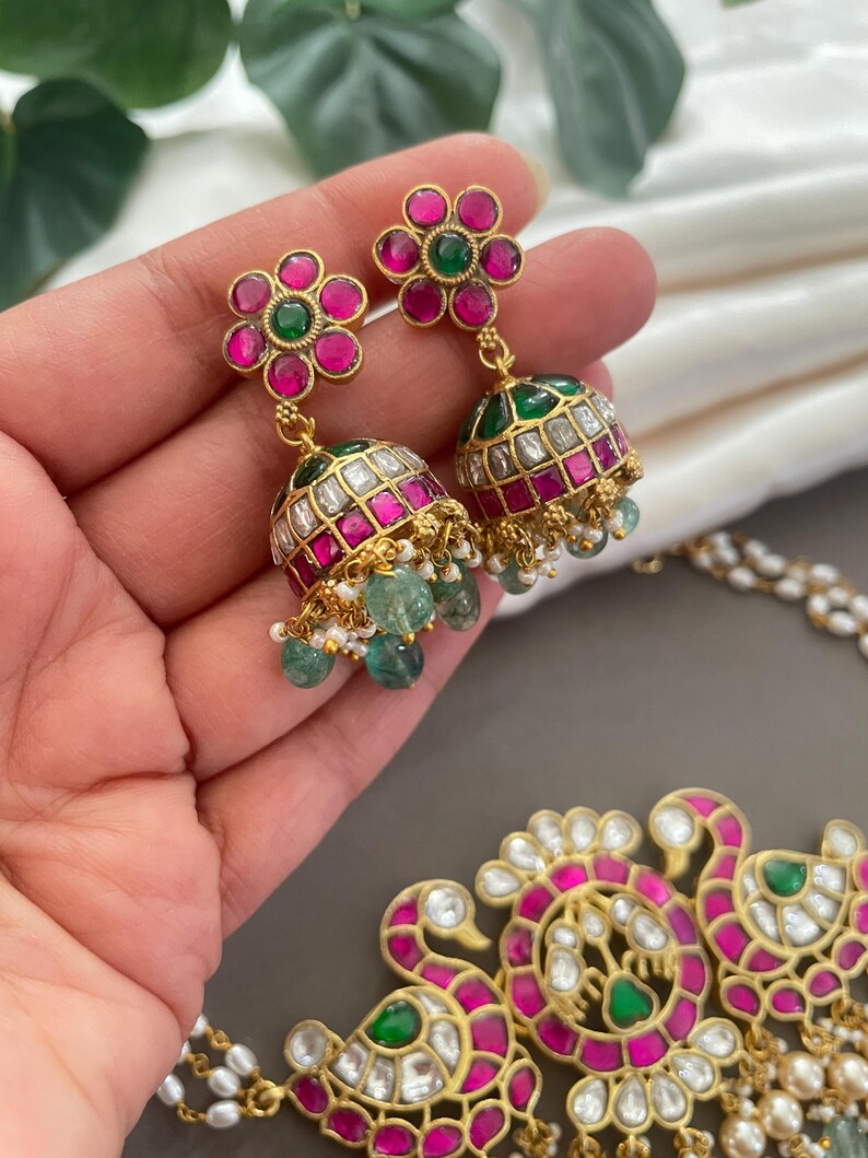 May include: A gold necklace with a pendant featuring two peacocks with pink and green gemstones. The necklace is paired with matching earrings with pink and green gemstones.