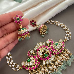 May include: A gold necklace with a pendant featuring two peacocks with pink and green gemstones. The necklace is adorned with white pearls and green beads.