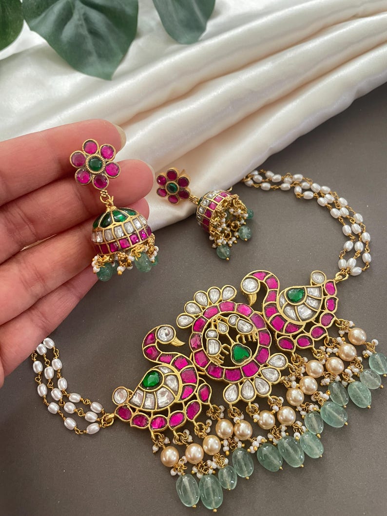 May include: A gold necklace with a pendant featuring two peacocks with pink and green gemstones. The necklace is adorned with white pearls and green beads.