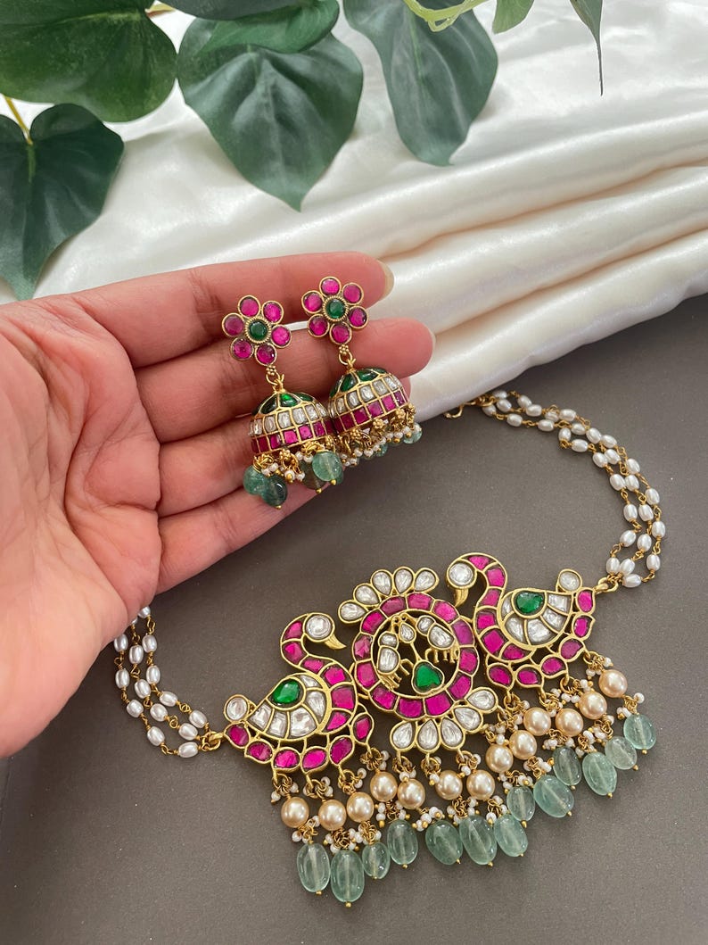 May include: A gold necklace with a peacock design featuring pink and green gemstones. The necklace has a pearl and green gemstone drop. Matching earrings with pink and green gemstones are also included.