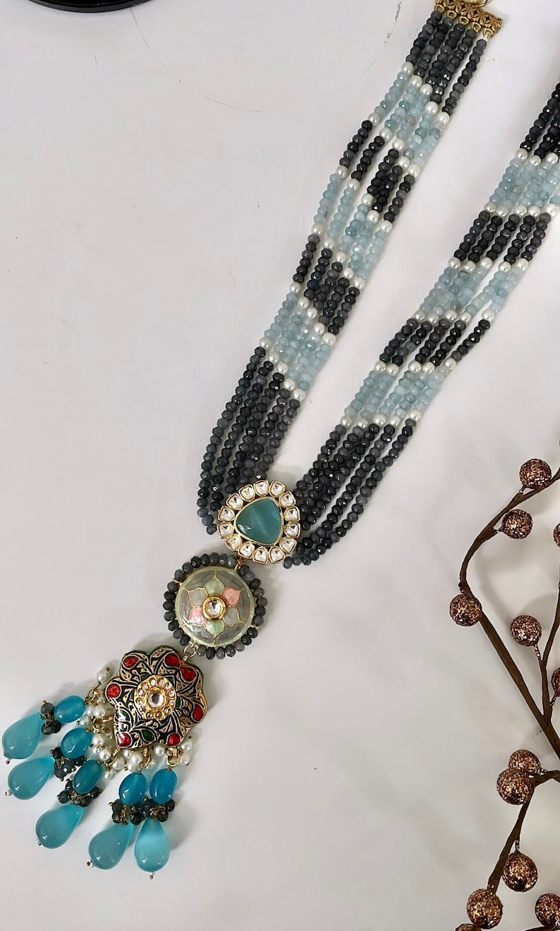 May include: A multi-strand necklace with black, white, and blue beads. The necklace has a large, ornate pendant with a green stone and a floral design.