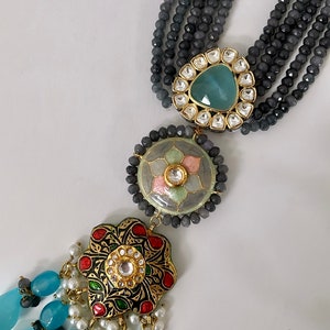 May include: A multi-strand necklace with gray, blue, and white beads. The necklace has a pendant with a blue, green, and gold design. The pendant is decorated with small, clear stones.