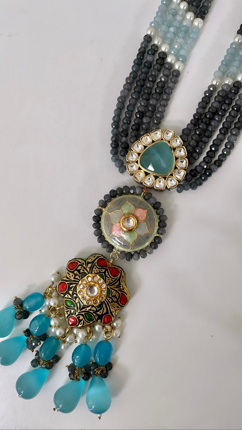 May include: A multi-strand necklace with gray, blue, and white beads. The necklace has a pendant with a blue, green, and gold design. The pendant is decorated with small, clear stones.