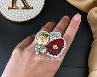 May include: A silver ring with a red, carved stone, a gold-toned circular design, and silver beads. The ring is on a hand.