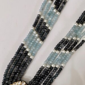May include: A multi-strand necklace with blue and gray beads, accented with white pearls. The necklace features a large, ornate pendant with a blue gemstone.