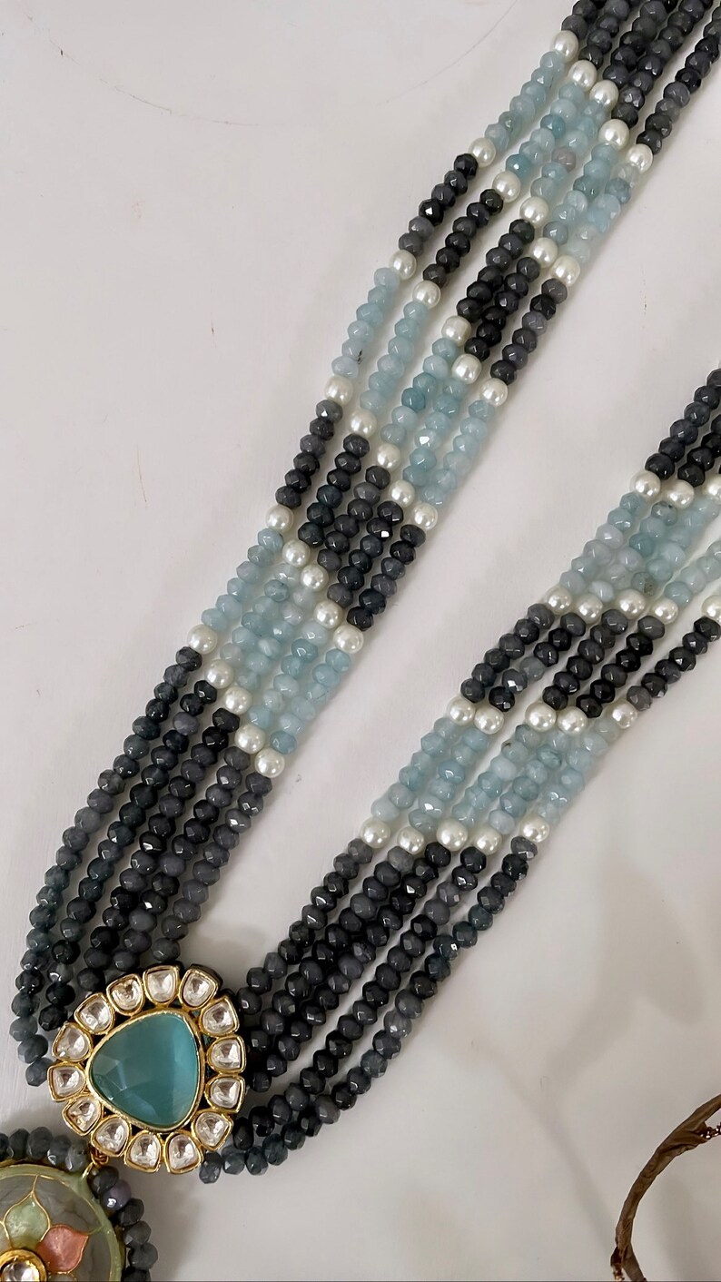 May include: A multi-strand necklace with blue and gray beads, accented with white pearls. The necklace features a large, ornate pendant with a blue gemstone.