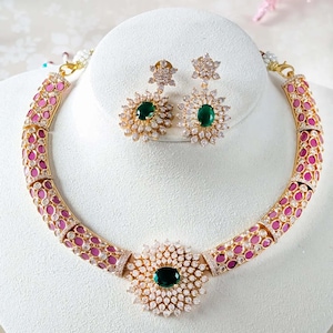 May include: A gold necklace with a large green stone surrounded by smaller white stones. The necklace is accented with pink stones. Matching earrings with green stones and white stones are included.