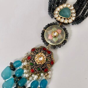 May include: A multi-strand necklace with blue, white, and black beads. The necklace has a large, ornate pendant with a floral design and a blue stone in the center. The pendant is attached to a smaller, round pendant with a floral design and a pink stone in the center.