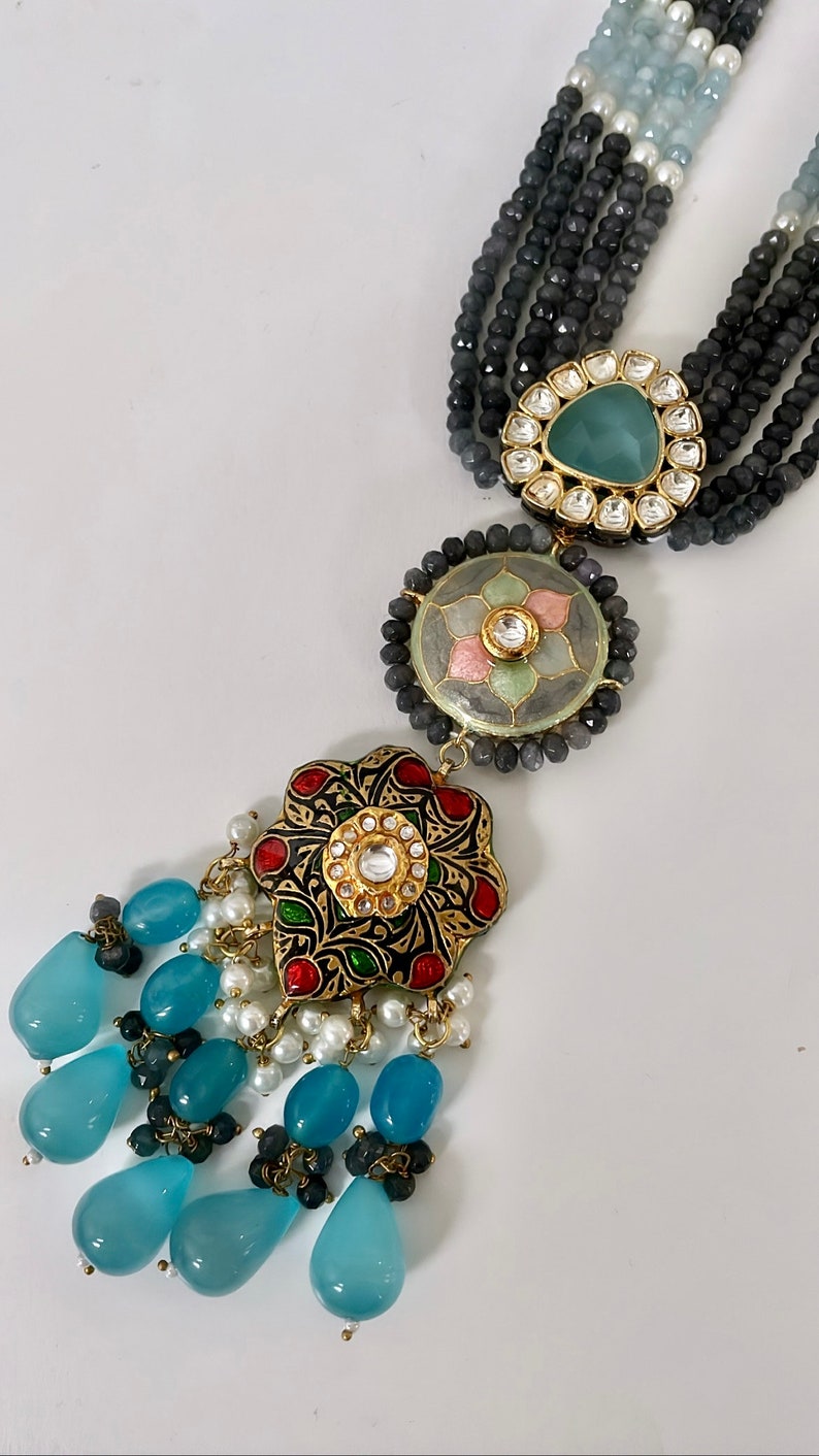 May include: A multi-strand necklace with blue, white, and black beads. The necklace has a large, ornate pendant with a floral design and a blue stone in the center. The pendant is attached to a smaller, round pendant with a floral design and a pink stone in the center.