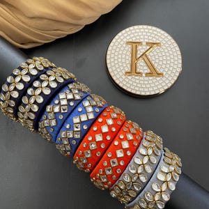 May include: A set of six silk thread bangles with intricate designs. The bangles are in shades of blue, orange, and gray, and are adorned with clear rhinestones. The bangles are stacked on top of each other, creating a visually appealing display.