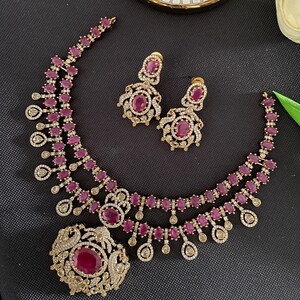 May include: A gold-coloured jewellery set comprising a necklace, earrings, and a brooch. The set is decorated with oval, deep red gemstones and small, clear crystals. The necklace has a double-strand design with teardrop accents.