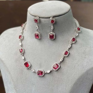 May include: A silver-toned necklace and earring set. The necklace and earrings feature square, ruby-red gemstones surrounded by small, clear stones. The earrings are dangling, and the necklace is displayed on a gray bust.