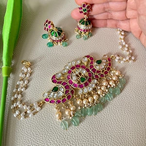 May include: A gold necklace with a large, ornate pendant featuring pink and green stones. The pendant is surrounded by a string of pearls and green stones. The necklace is paired with matching earrings.
