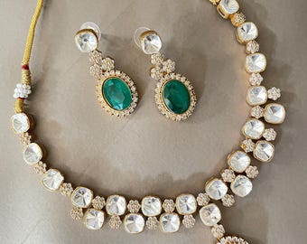 May include: A gold-toned necklace and earring set. The necklace features clear, square-cut stones and a large emerald-green oval pendant. The matching earrings have a similar design with emerald-green oval stones. The set is on a neutral background.