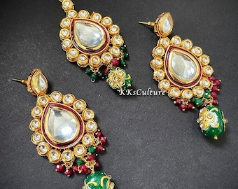 May include: A gold-tone Indian bridal jewellery set with a teardrop-shaped pendant and matching earrings. The set features clear stones, red beads, and green accents.