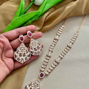May include: A gold necklace and earrings set with a pink stone pendant and matching earrings. The necklace features a delicate chain with small white stones and a large, ornate pendant with a pink stone in the center. The earrings are similarly designed with a pink stone and small white stones.