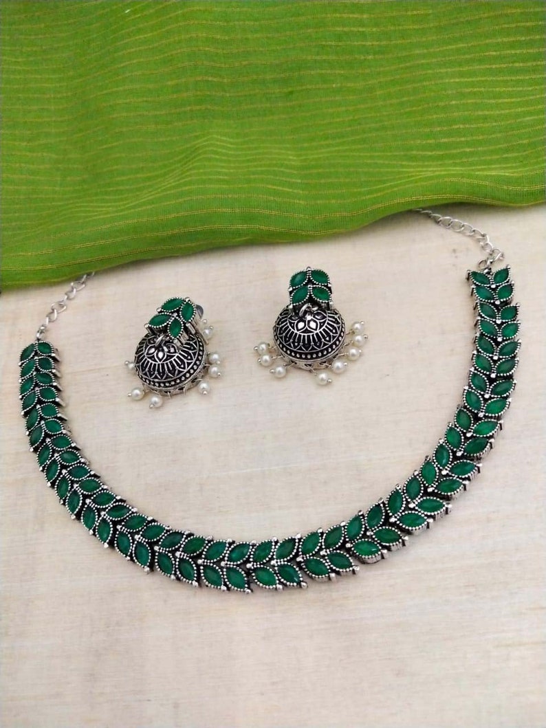 May include: A silver necklace with green gemstone leaves and matching earrings. The necklace and earrings have a traditional Indian design.