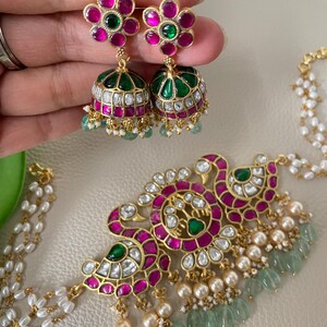 May include: A gold necklace with a peacock design, featuring pink and green gemstones, white pearls, and green beads. The necklace is paired with matching earrings with pink and green gemstones.