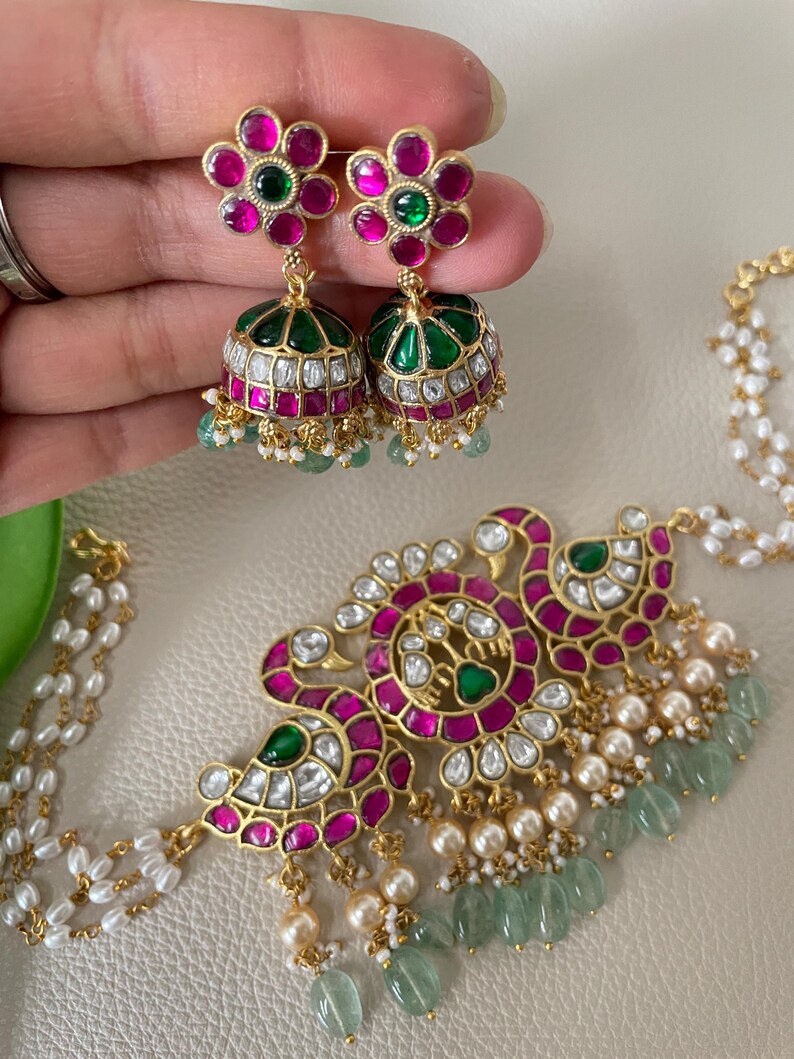 May include: A gold necklace with a peacock design, featuring pink and green gemstones, white pearls, and green beads. The necklace is paired with matching earrings with pink and green gemstones.